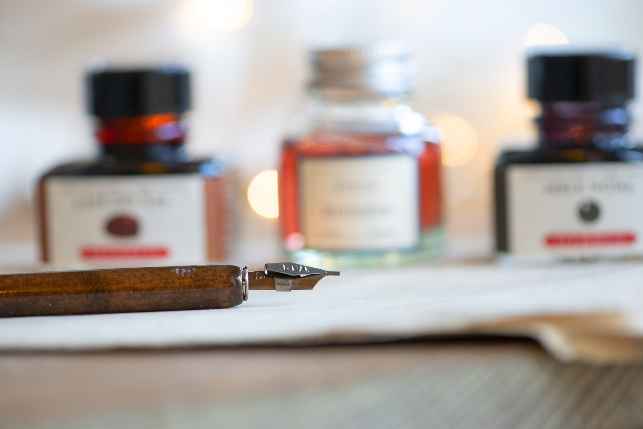 Plume d’écriture posée sur une feuille de papier, avec des flacons d’encre flous en arrière-plan.