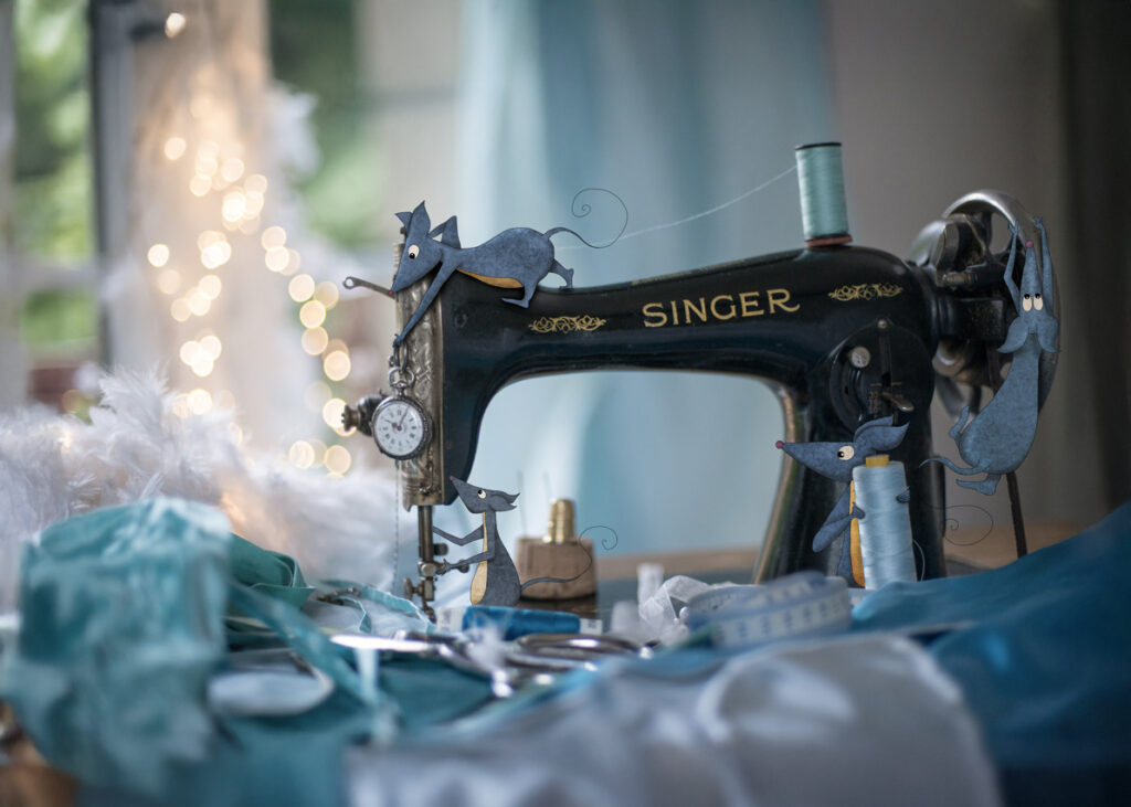 Photographie d’une machine à coudre transformée en décor miniature, avec des souris illustrées s’activant autour du fil et de l’aiguille pour confectionner une robe.