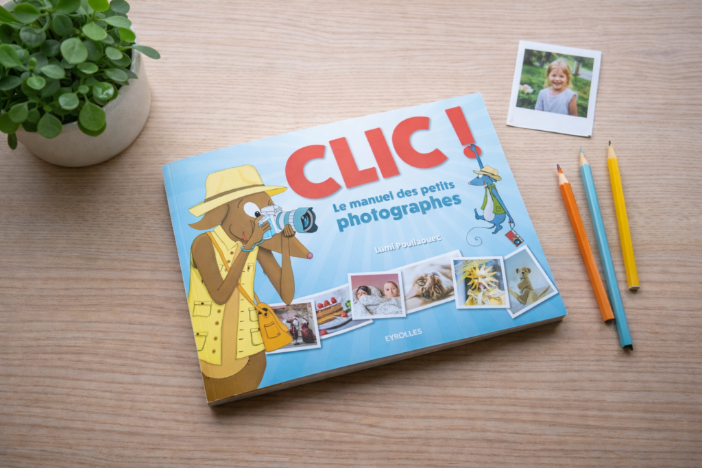 Livre jeunesse « CLIC ! Le manuel des petits photographes » posé sur une table en bois clair, décoré d’une plante verte, de crayons de couleur et d’un polaroid.