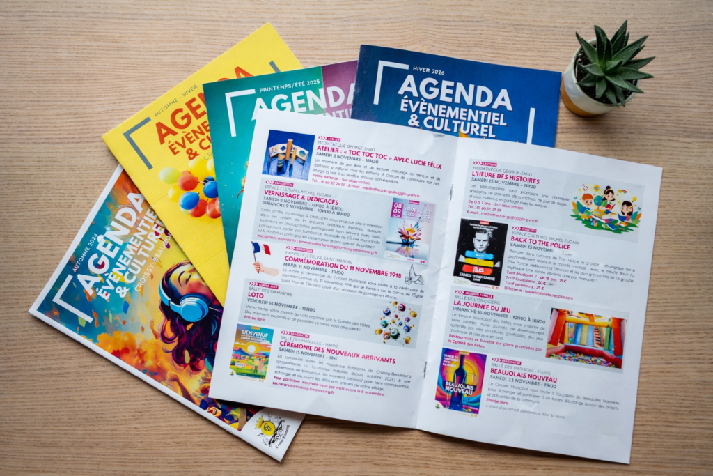 Plusieurs numéros de l’Agenda événementiel et culturel disposés sur une table en bois, avec une petite plante verte à côté.