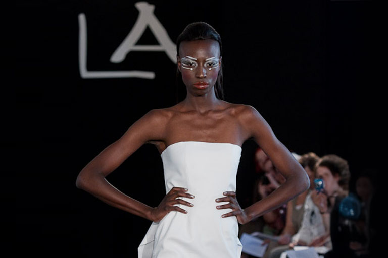 Mannequin portant une robe blanche bustier, mains sur les hanches, défilant sur un podium lors d’un défilé de mode avec un fond noir et des spectateurs en arrière-plan.