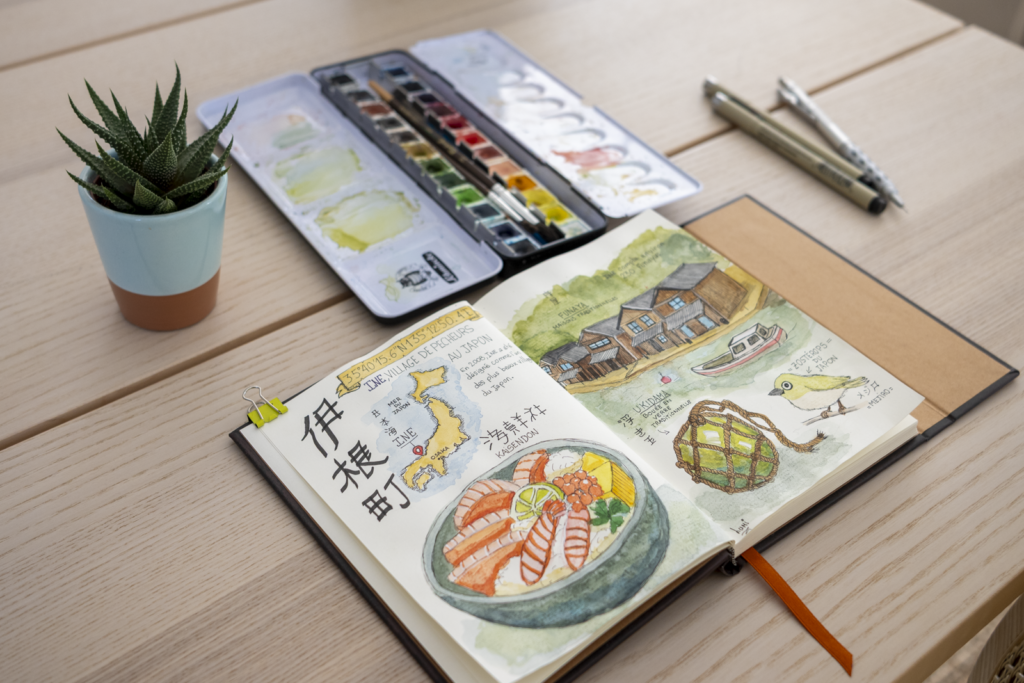 Carnet de voyage illustré ouvert sur une page dédiée à Ine au Japon, avec aquarelles d’un kaisendon, des maisons funaya, une bouée en verre et une carte du Japon, posé sur un bureau avec palette d’aquarelle et stylos.
